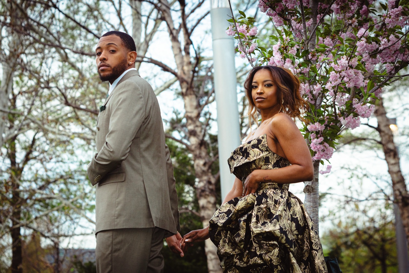 Kimberly in a gold brocade gown and Jelani in a sage suit, holding hands under pink cherry blossoms