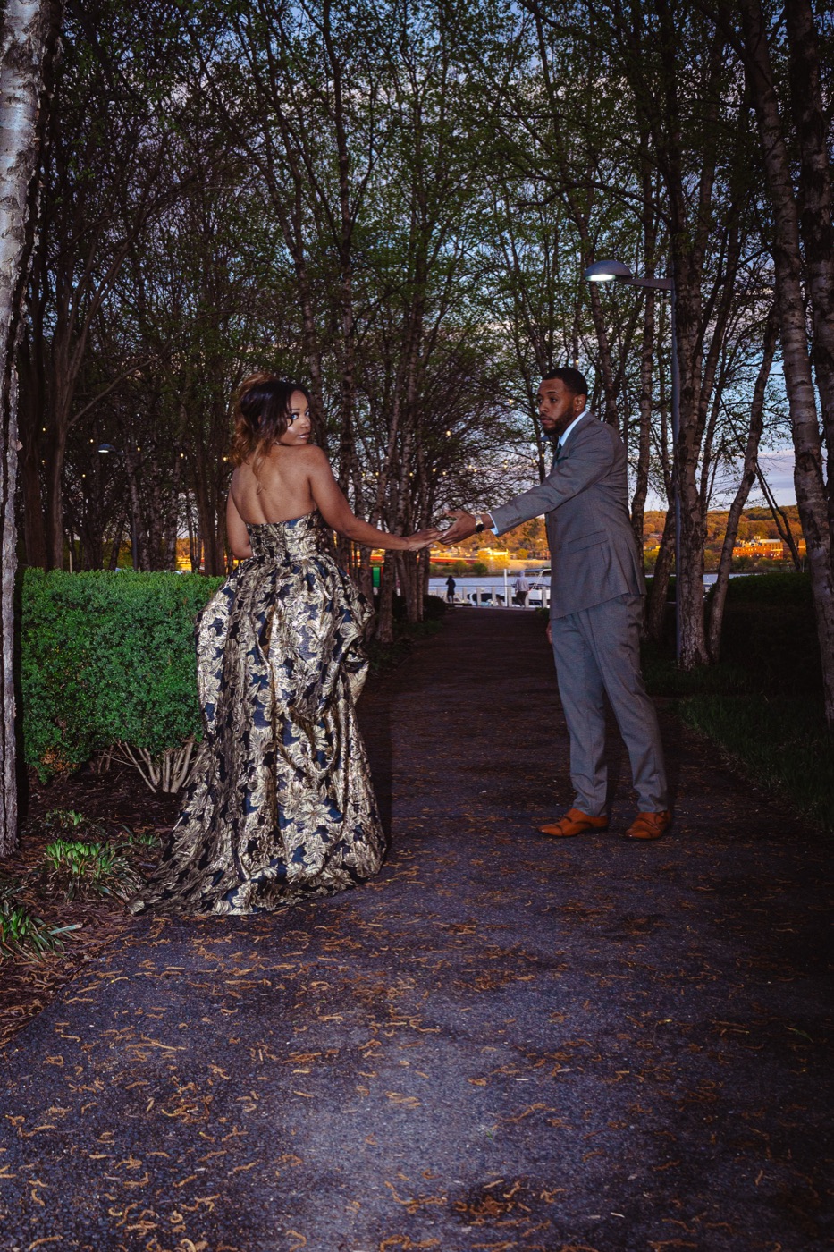 Jelani reaching for Kimberly's hand on a tree-lined path by the water at dusk
