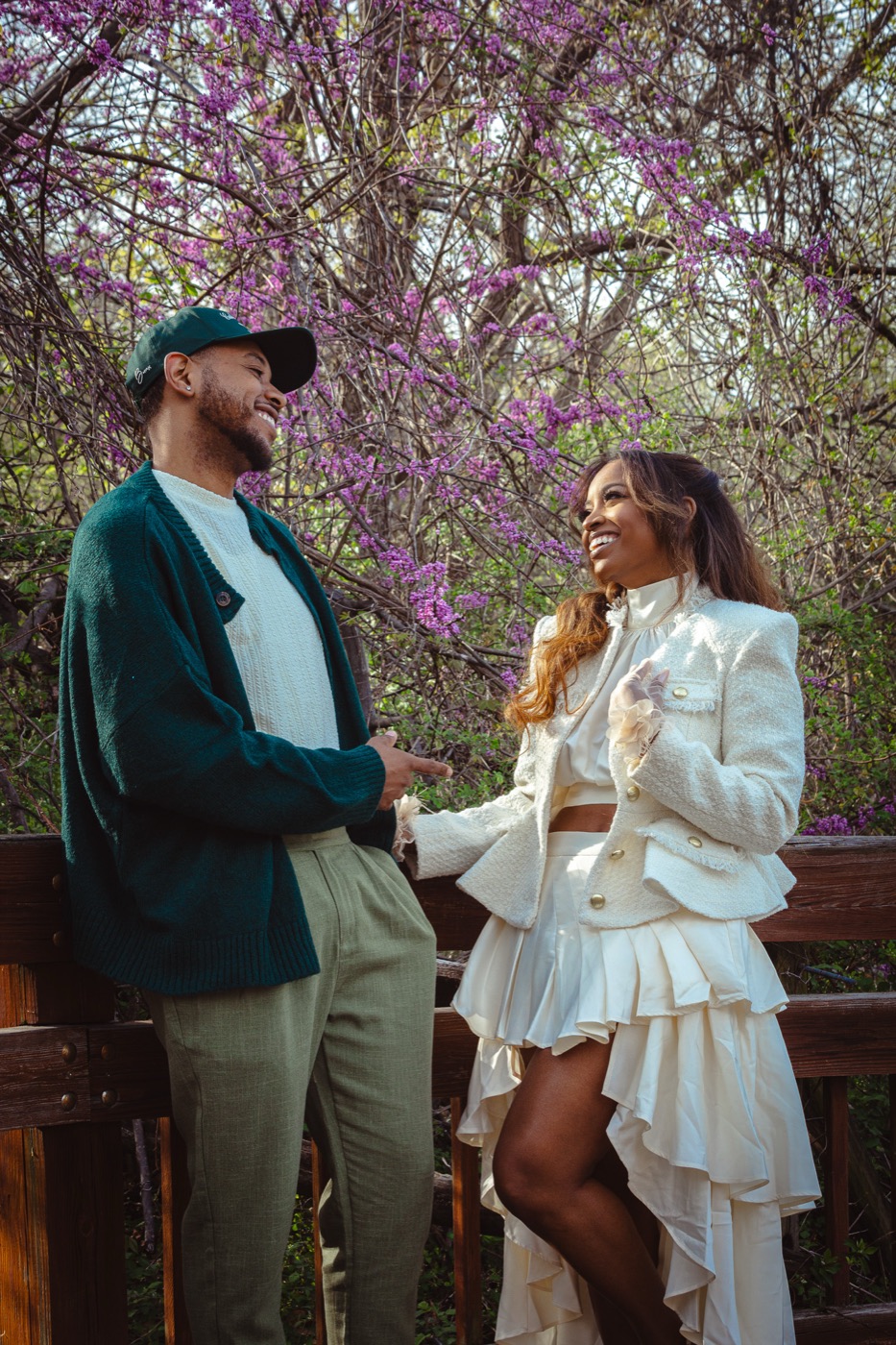 Kimberly and Jelani laughing together on a wooden bridge under purple blossoms