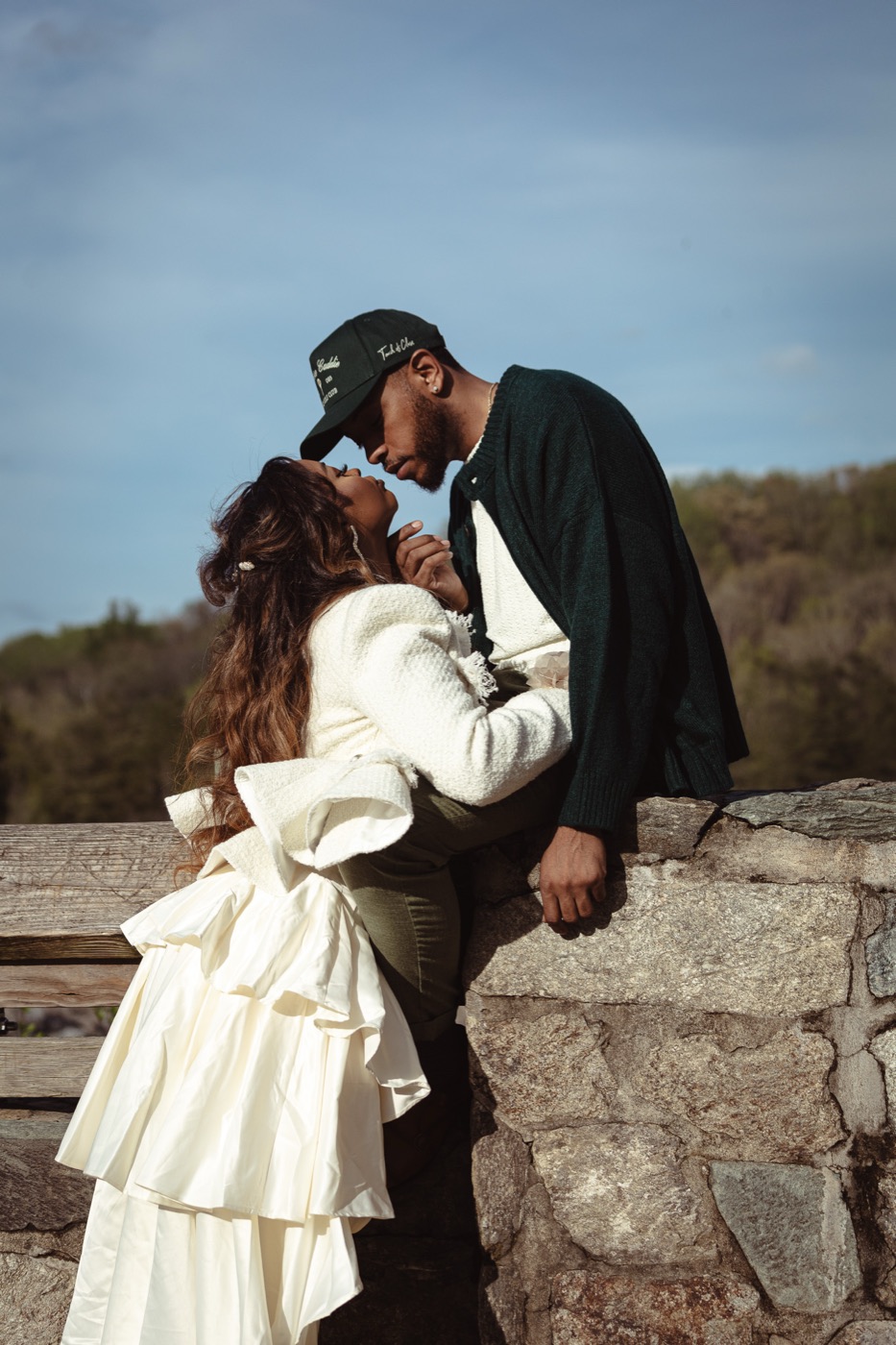 Jelani leaning in to kiss Kimberly against a stone wall under a blue sky
